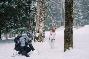 Little girl stands in a snowy forest near her mother making a snowman while squatting photo