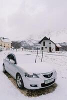 Snow-covered car with raised windshield wipers stands near a cottage photo