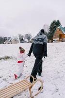 Mother with a sled on a rope and a little girl climb a hill holding hands. Back view photo