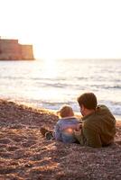 Dad is lying on the beach next to a little girl sitting and looking at the sea. Back view photo