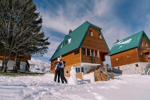 Mom kisses dad with little girl on his shoulders standing next to snowman near wooden chalet photo