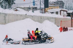 Dad and children ride on a snowmobile with a sled attached to the back along a snowy mountain photo