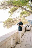 fotógrafo con un cámara en su manos soportes en un terraza con vista a el mar debajo un árbol foto