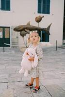 pequeño niña soportes abrazando un rosado juguete Conejo en el patio de un antiguo edificio y mira arriba foto