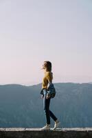 joven mujer en Gafas de sol soportes en un Roca bordillo alto en el montañas. lado ver foto