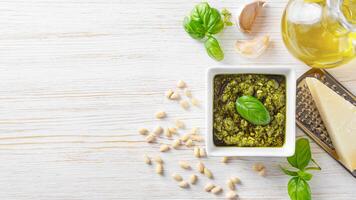 Homemade pesto sauce in small square jar and ingredients for pasta on white wooden background photo