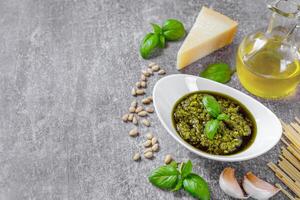 Homemade pesto sauce in small round white jar and ingredients on gray background, copy space photo