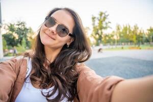 Young smiling woman in trendy summer clothes. Happy model posing in the street at sunny day. Makes pov selfie in sunglasses. High quality photo