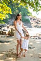 joven mujer madre con un pequeño hija en blanco vestidos teniendo un divertido en costa en el sombra de arboles y palmas alto calidad foto