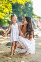 joven mujer madre con un pequeño hija en blanco vestidos en costa en el sombra de arboles y palmas alto calidad foto