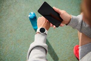 Male athlete using smartphone with black mockup screen, checking mobile app on his wrist watch, after workout outdoor photo