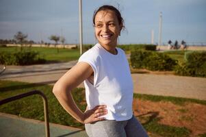 Young happy fitness woman, athlete smiling after running, looking away, relaxing on sports ground after outdoor workout photo