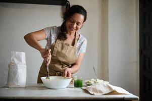 Authentic pretty woman housewife in beige apron, using wooden spoon, mixing spinach water with whole grain flour for making dough, standing at marble kitchen table in the countryside rural house photo