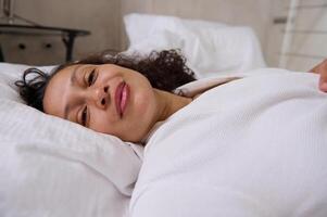 Close-up portrait of curly haired mixed race young woman smiling looking at camera, lying on the bed at home bedchamber photo