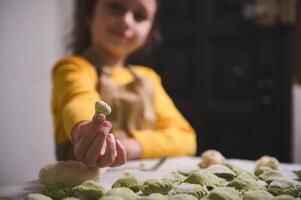 Close-up hand of blurred child girl, little chef in beige apron, holds out at camera a homemade dumpling, smiles looking at camera, standing at floured table with molded ravioli, pelmeni, vareniki photo