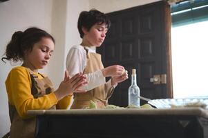 Kids modeling dumplings, standing at floured table at home kitchen interior. Adorable brother and sister preparing a family dinner together, sculpting varennyki according to traditional recipe photo