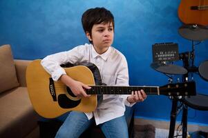 adolescente chico jugando acústico guitarra a hogar, sentado en un silla en contra musical instrumentos y azul pared antecedentes foto