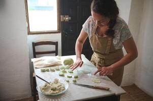 Housewife making dumplings at home kitchen. Pretty woman in beige apron rolling out the dough with a glass bottle as a rolling pin photo