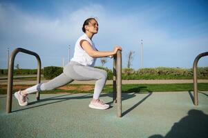 determinado Deportes mujer en blanco camiseta y gris polainas, calentamiento arriba, haciendo estocadas para extensión pierna músculos. foto