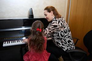 Inspired happy woman pianist, music teacher performing melody on piano forte, explaining piano lesson to a child girl, sitting nearby on a stool. Musical education and talent development in progress photo