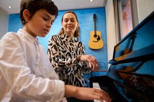 Teenager student boy sitting at piano near her teacher, having piano lesson at home. Different musical instruments against a blue color wall on the background. People. Education. Lifestyle photo