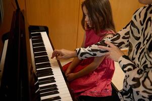pequeño niño niña en elegante rojo vestido, tomando piano lección, apasionadamente jugando el llaves debajo su del maestro guía, sensación el ritmo de música. musical educación y talento desarrollo en Progreso foto