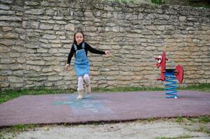 Sporty kid girl plays hopscotch, takes turns jumping over squares marked on playground. Childhood and healthy lifestyle photo