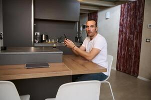 Smiling young man looking at camera while using smartphone for online shopping, sitting at table at home photo