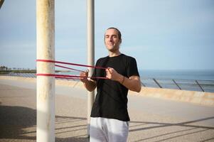 Young athletic man working out on promenade, doing shoulder press using resistance band. Active healthy lifestyle. Sport photo