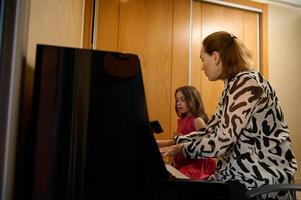 woman pianist holding hands on piano keyboard, touching black and white keys, preforming classical melody during while giving a music lesson to her little student girl. photo