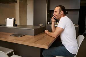 joven hombre sonriente mientras hablando en móvil teléfono, recepción llamada desde amigos, sentado a mesa en el hogar interior foto