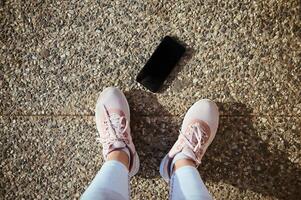 deportista punto de vista piernas y pies de hembra atleta en rosado zapatillas y teléfono inteligente con Bosquejo negro pantalla en el asfalto foto