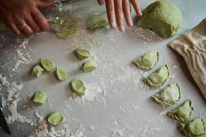 Close-up top view of woman cook chef in beige apron, standing at kitchen table with ingredients, rolling out the dough with a wine bottle as rolling pin, dumplings at home kitchen. Copy space photo