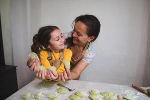 Smiling mother and daughter looking at each other, holding sculpted homemade dumplings or Ukrainian varenyky, standing together at floured kitchen table, against white wall background photo