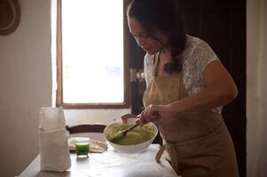 Gorgeous woman chef, housewife in beige apron, standing by kitchen table, kneading dough with wooden spoon, preparing traditional ravioli according to traditional Italian recipe, in rural environment photo