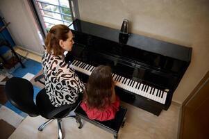 View from above of a music teacher, pianist musician mother giving a piano lesson to her daughter, a cute little kid girl dressed in elegant red dress, sitting together at piano and performing melody photo