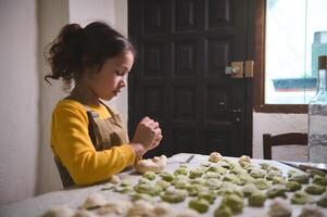 confidente retrato de linda pequeño niña en beige delantal, en pie a cocina mesa con moldeado albóndigas, Relleno el masa y moldura Ravioles, Ayudar su mamá en culinario, preparando cena solo foto