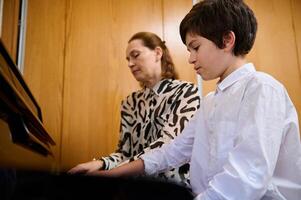 Confident teenage boy pianist musician creating the rhythm of melody, touching white and black keys of piano while playing pianoforte with his music teacher , sitting nearby on the blurred background photo
