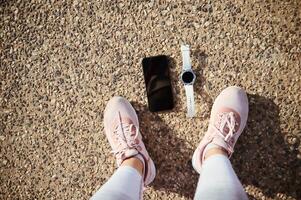 Top view Legs and feet of female athlete in pink sneakers, smart wristwatch and smartphone with mockup screen on asphalt photo