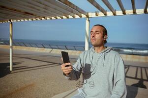 Caucasian athlete man wearing earphones, listening to music while resting after workout against Atlantic beach backdrop photo