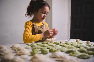 adorable caucásico pequeño niño chica, en amarillo suéter y beige cocinero delantal, en pie a cocina mesa con moldeado albóndigas, preparando familia cena. Cocinando clase para niños foto