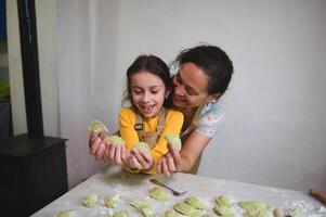 mamá y hija tener divertido mientras Cocinando albóndigas, en pie juntos a enharinado cocina mesa foto