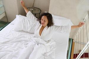 View from above young woman stretching, lying in comfortable bed on soft pillows and white bed sheets, smiling at camera photo