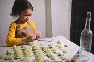 pequeño niña moldura albóndigas, en pie a enharinado cocina mesa. niño niña Ayudar su mamá Cocinando cena a hogar foto