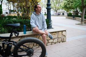 caucásico joven hombre sentado en un banco en el ciudad, relajante después ciclismo en eléctrico bicicleta. un bicicleta eléctrica en el primer plano foto