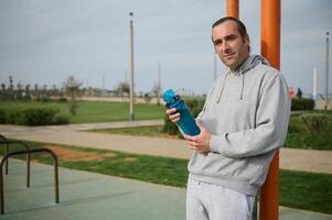 Determined young athlete holding water bottle, resting after outdoor workout on an urban sportsground, looking at camera photo