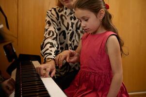 confidente retrato de un hermosa caucásico pequeño niño niña en elegante rojo vestido, aprendizaje jugando en pianoforte. hembra pianista profesor explicando lección, sentado juntos a grandioso piano foto