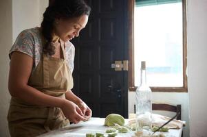 bonito ama de casa en beige delantal, Cocinando Ravioles empanadillas con machacado patatas,de pie a cocina mesa con ingredientes en rústico acogedor Doméstico cocina. gente, culinario, rutina y pasatiempo concepto foto