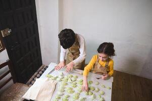 Kids modeling dumplings, standing at floured table at home kitchen interior. Top view boy and girl preparing a family dinner together, sculpting green ravioli according to Italian traditional recipe photo