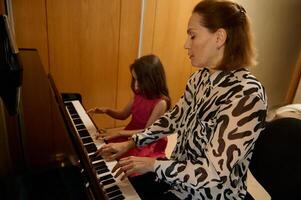 auténtico retrato de un joven adulto madre y hija sentado a grandioso piano, ejecutando clásico melodía, tocar blanco y negro teclado de el piano forte, jugando piano, canto canción foto